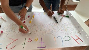Zwei Personen malen etwas mit Buntstiften auf ein großes Plakat unter der Überschrift "Holi Fest 23" | © Caritas Don Bosco Berufsfachschule für Kinderpflege