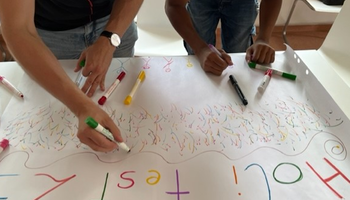 Zwei Personen malen etwas mit Buntstiften auf ein großes Plakat unter der Überschrift "Holi Fest 23" | © Caritas Don Bosco Berufsfachschule für Kinderpflege